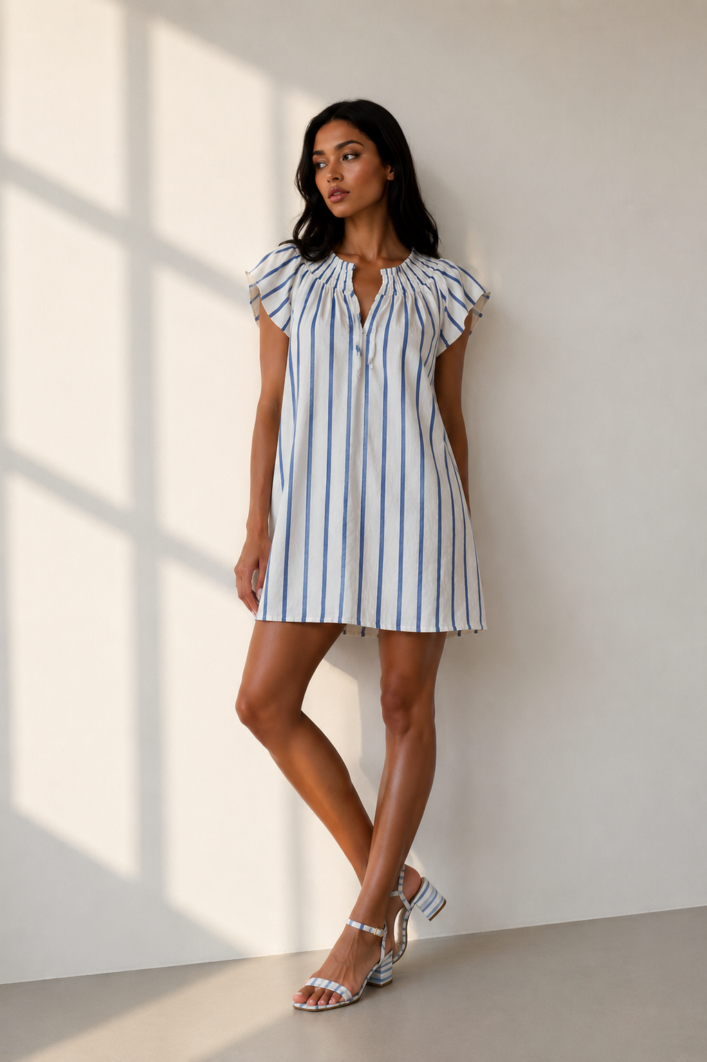 Model wearing the DayTrip Stripe blue and white striped mini dress by Sadie & Sage, standing in a sunlit room.