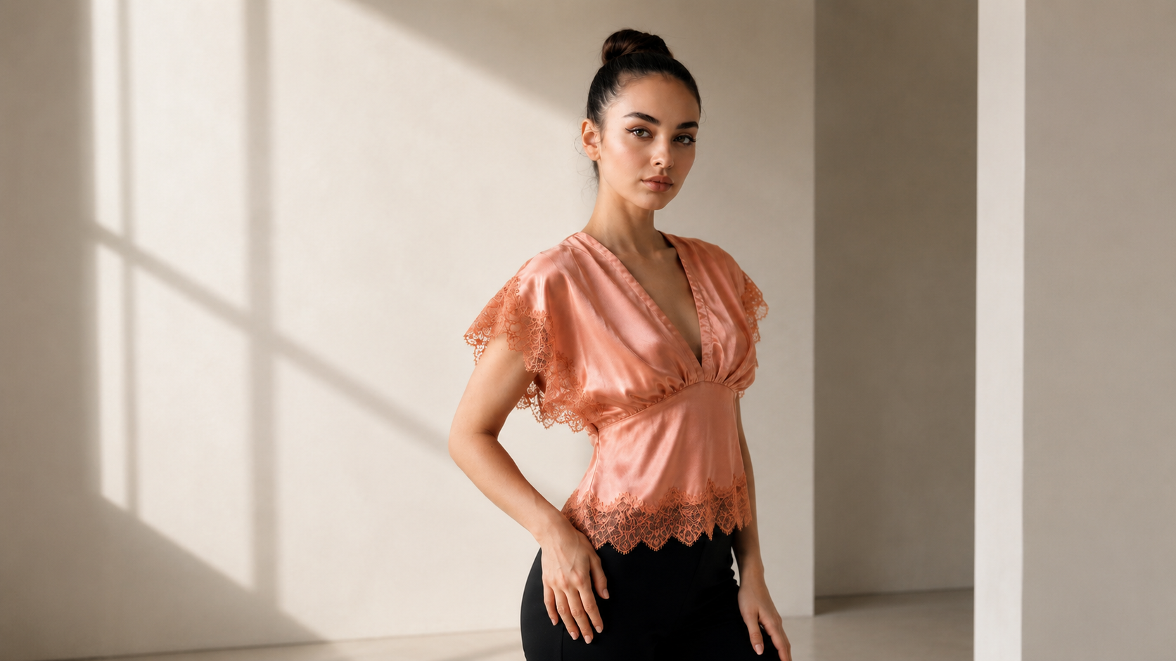 Wide shot of a model wearing the Satin Lace Top and black pants, standing in a sunlit room.